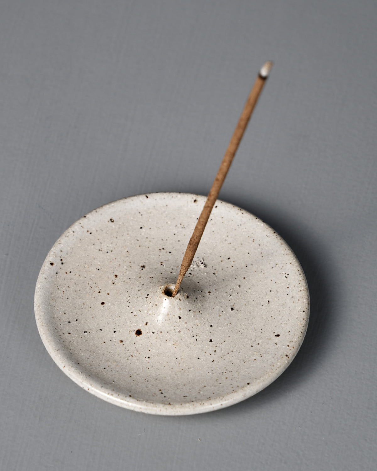 The Incense Holder – Speckled White, a handmade stoneware piece from Pōneke Wellington, displays its unique speckled finish while holding a single lit incense stick on a gray surface.