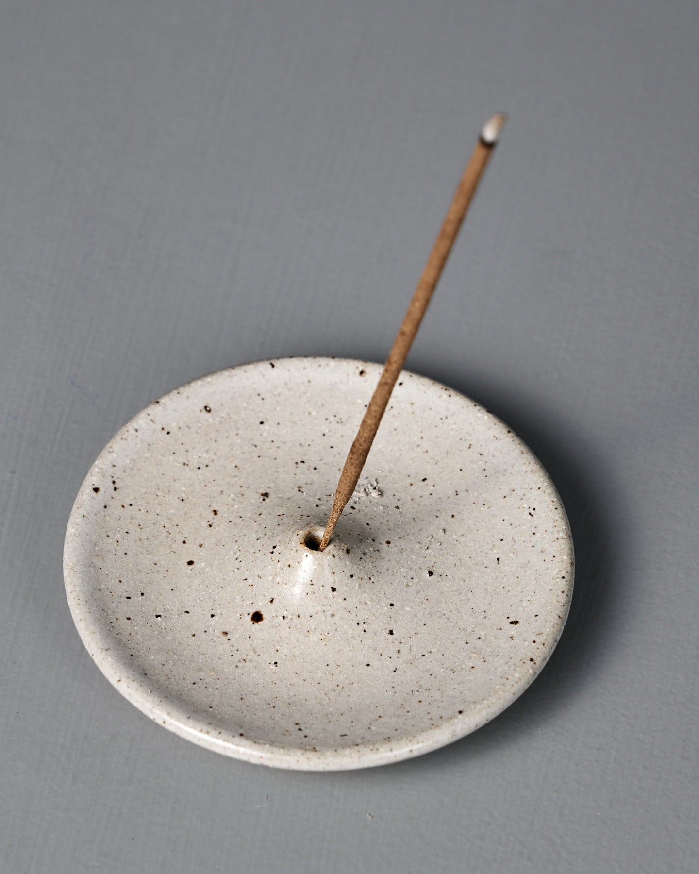 A lit incense stick stands upright in the Incense Holder – Speckled White, with a thin trail of smoke rising against a plain gray background.