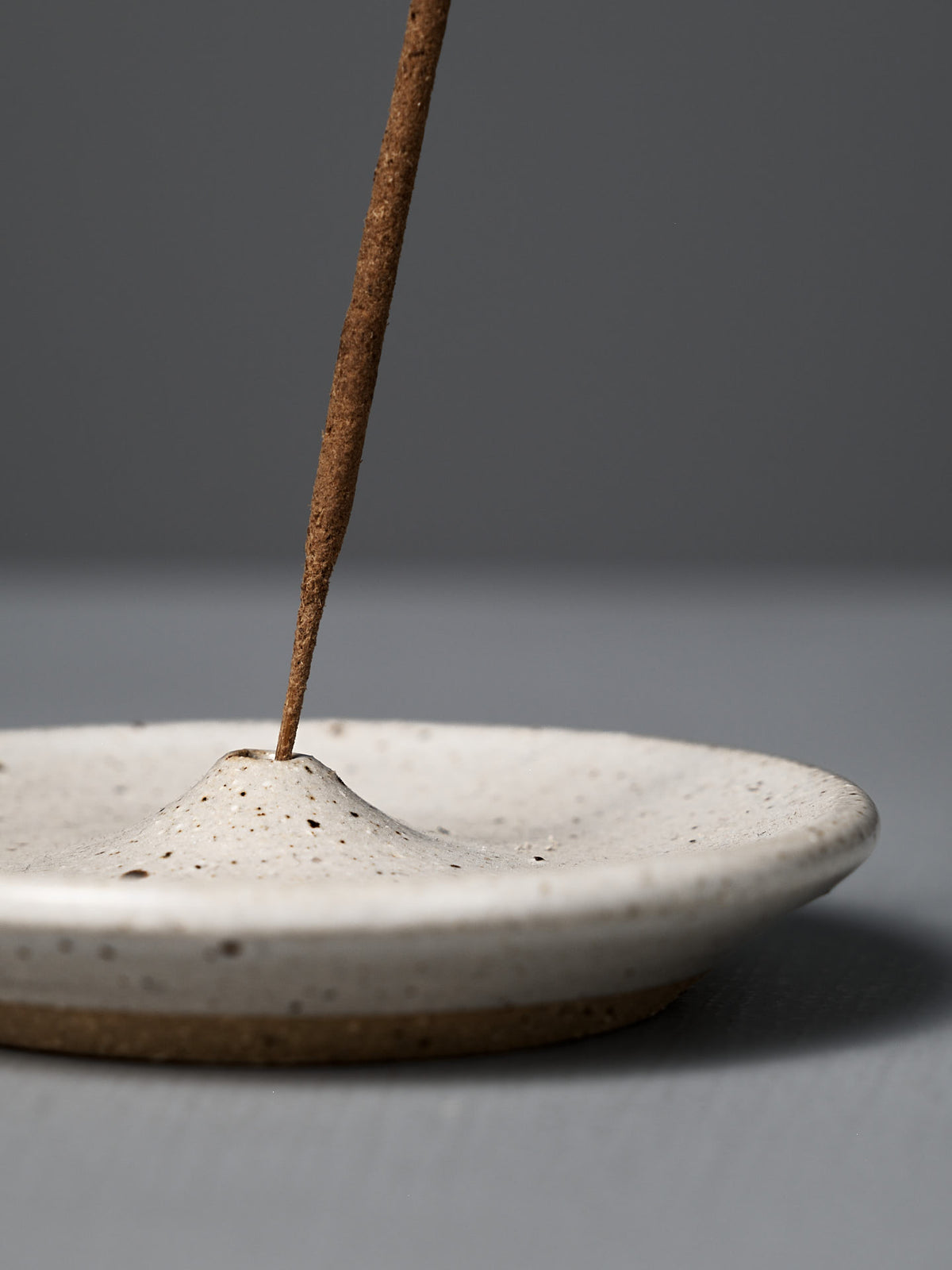 A brown incense stick stands upright in the handmade Incense Holder – Speckled White, featuring a central mound, set against a gray background. Perfect for adding a touch of Wellington-inspired style to your space.