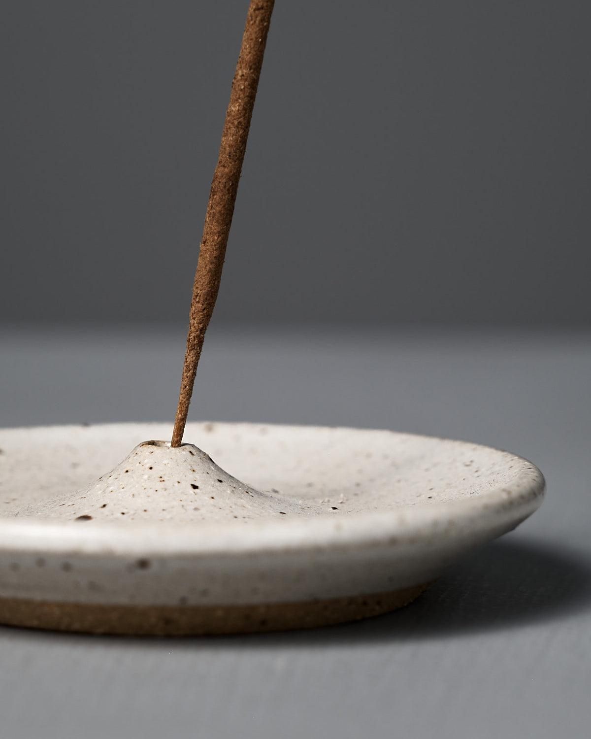A brown incense stick stands upright in the handmade Incense Holder – Speckled White, featuring a central mound, set against a gray background. Perfect for adding a touch of Wellington-inspired style to your space.