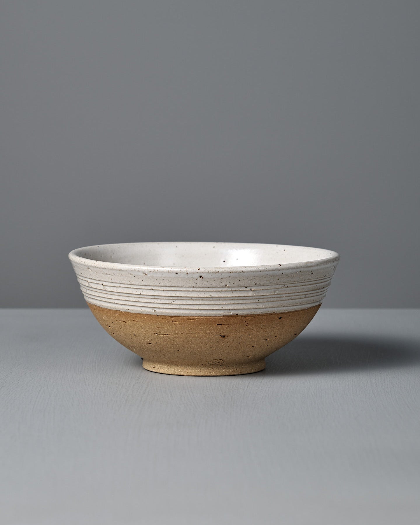 The Ridge Line Bowl, handmade with a matte speckled white glaze on the upper half, unglazed tan lower half, and horizontal textured lines, is displayed on a gray surface against a plain gray background.