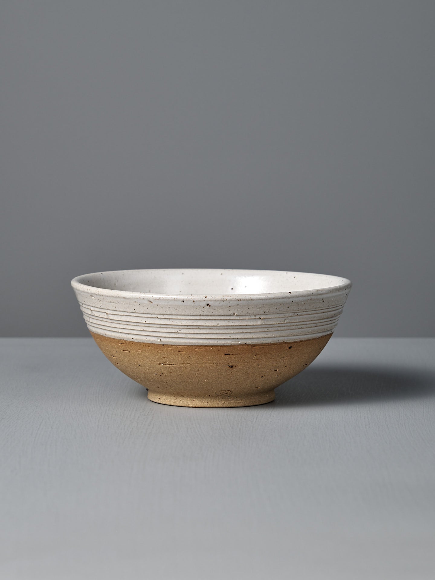 The Ridge Line Bowl, handmade with a matte speckled white glaze on the upper half, unglazed tan lower half, and horizontal textured lines, is displayed on a gray surface against a plain gray background.