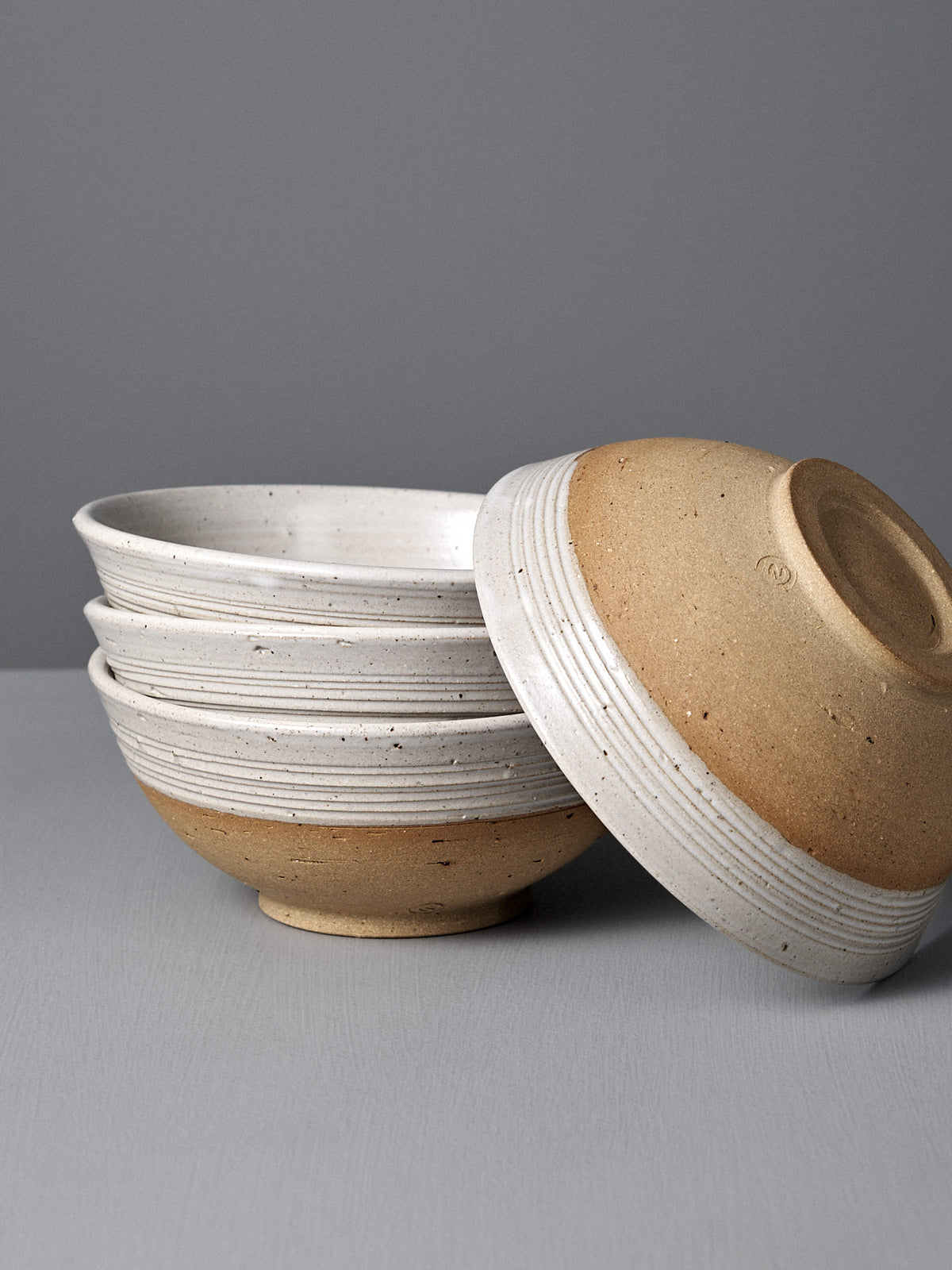 Four Ridge Line Bowls with a speckled white glaze are stacked on a gray surface, one handmade bowl resting upside down on top—a stylish touch of Wellington pottery for your table.