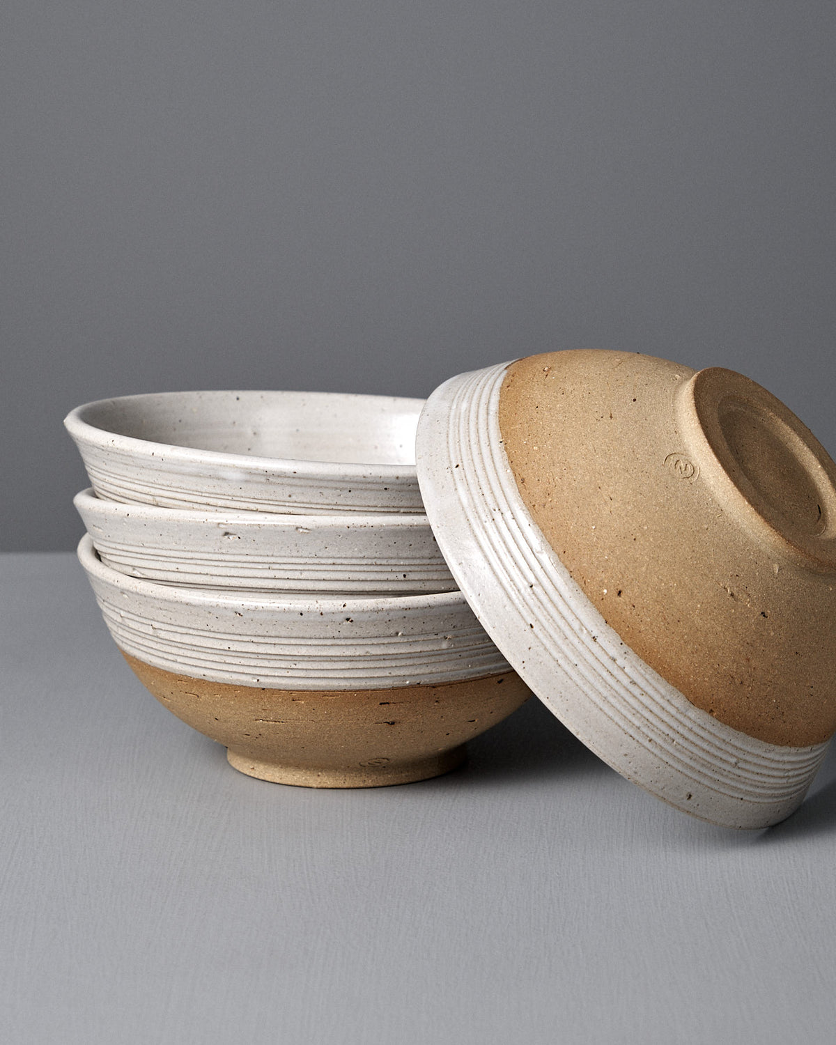 Four Ridge Line Bowls with a speckled white glaze are stacked on a gray surface, one handmade bowl resting upside down on top—a stylish touch of Wellington pottery for your table.