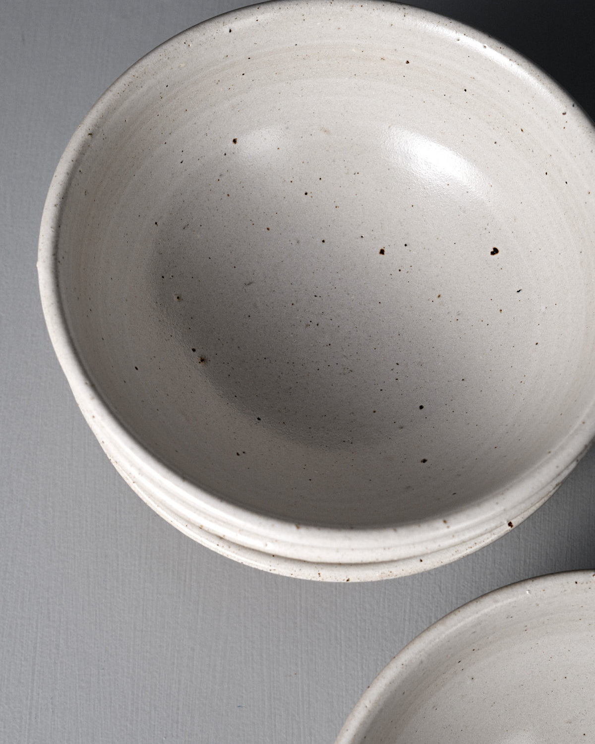 Three stacked Ridge Line Bowls with a speckled white glaze and matte finish rest on a gray surface, photographed from above—a beautiful example of Wellington pottery craftsmanship.