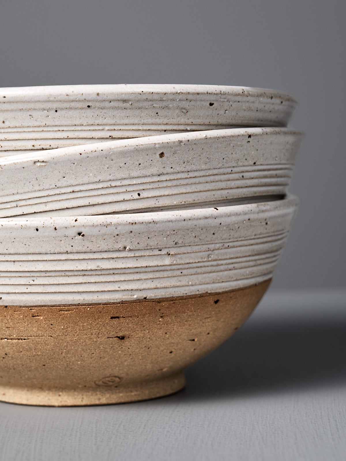 Three stacked Ridge Line Bowls feature a rustic, speckled white glaze on the top half and unglazed brown clay on the bottom, inspired by Wellington pottery, displayed against a plain gray background.