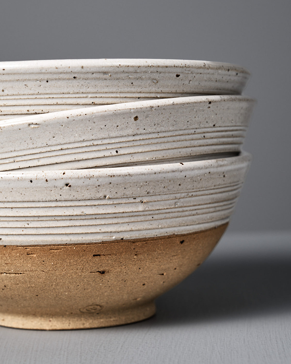 Three stacked Ridge Line Bowls feature a rustic, speckled white glaze on the top half and unglazed brown clay on the bottom, inspired by Wellington pottery, displayed against a plain gray background.