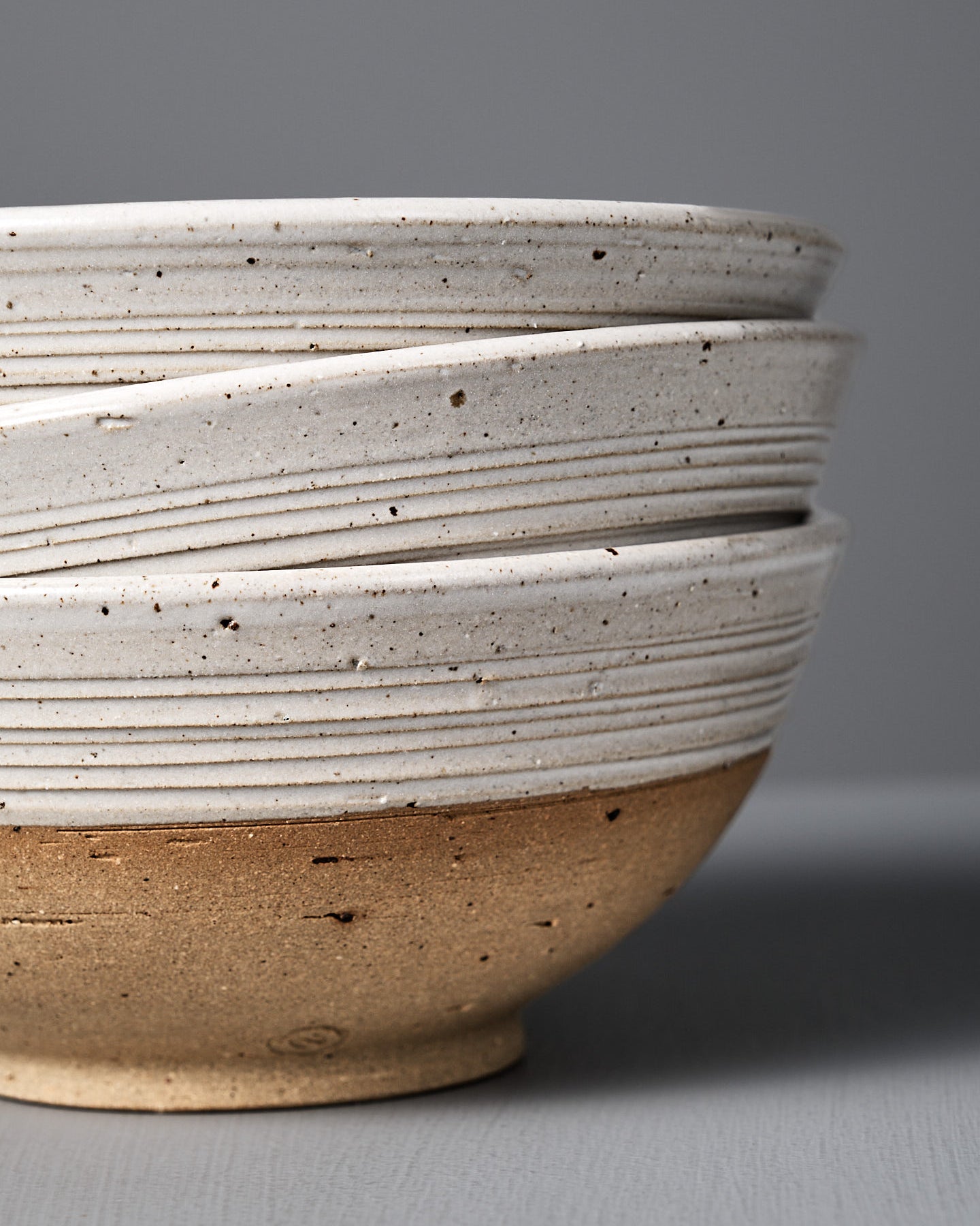 The Ridge Line Bowl, handmade with a matte speckled white glaze on the upper half, unglazed tan lower half, and horizontal textured lines, is displayed on a gray surface against a plain gray background.