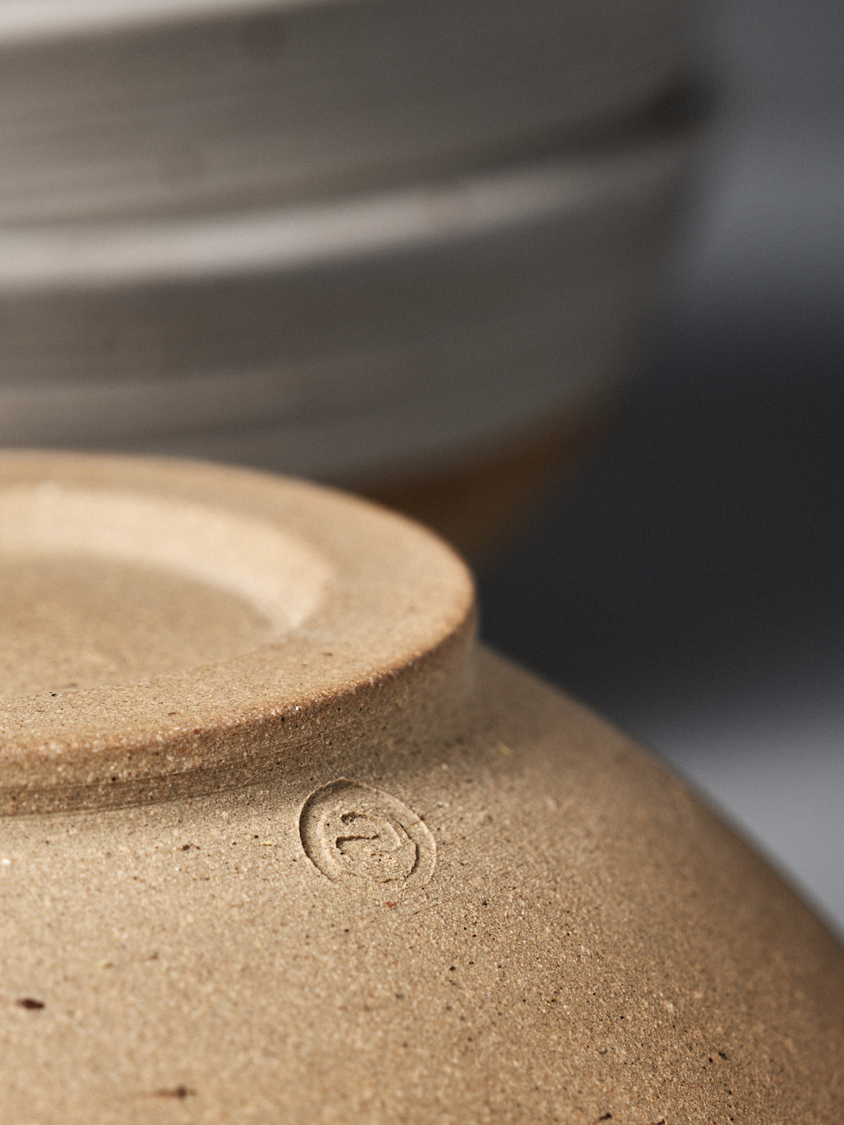 Close-up of the base of the Ridge Line Bowl, handmade with a speckled white glaze and featuring a small maker’s mark, with another ceramic piece softly blurred in the background.