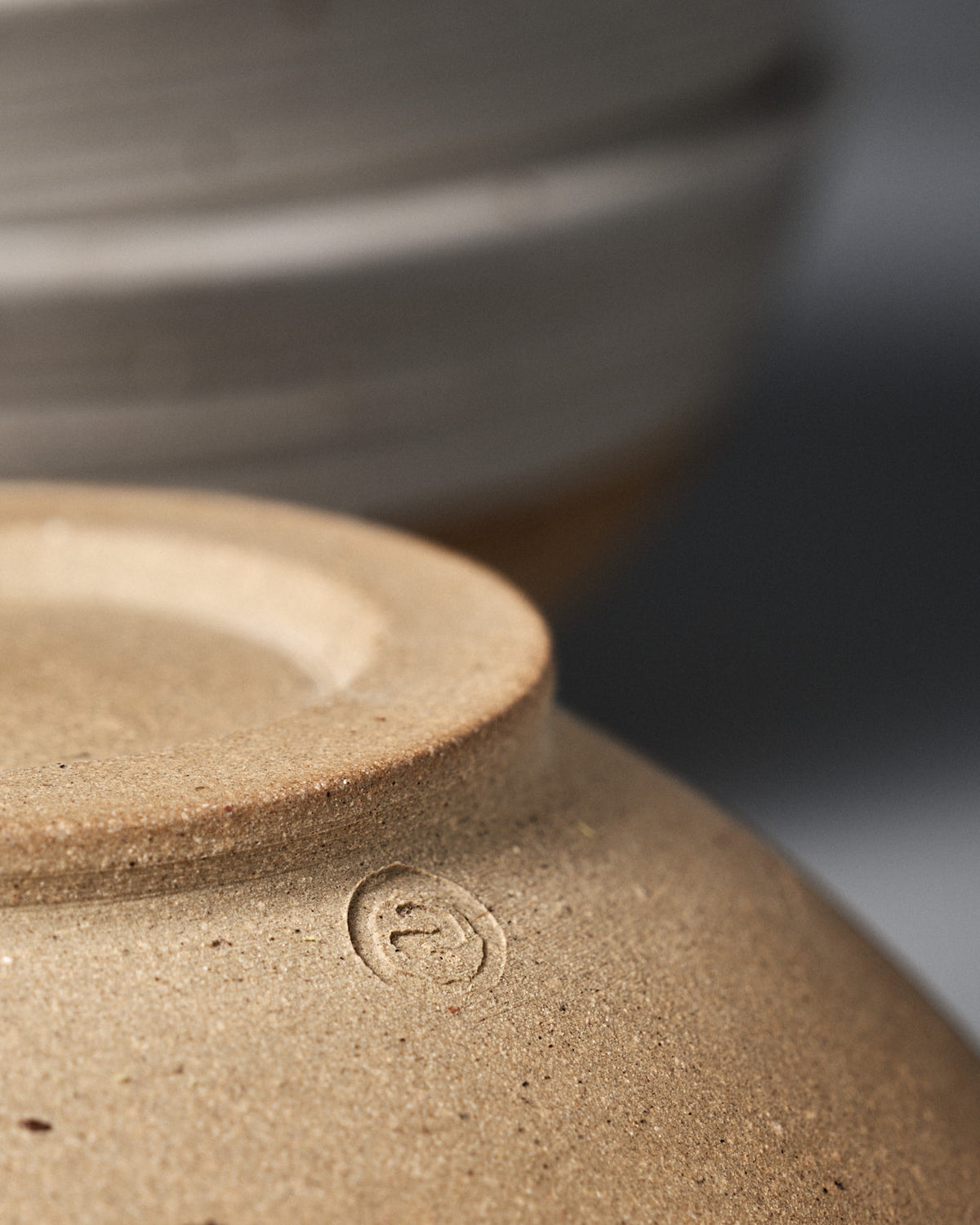 Close-up of the base of the Ridge Line Bowl, handmade with a speckled white glaze and featuring a small maker’s mark, with another ceramic piece softly blurred in the background.