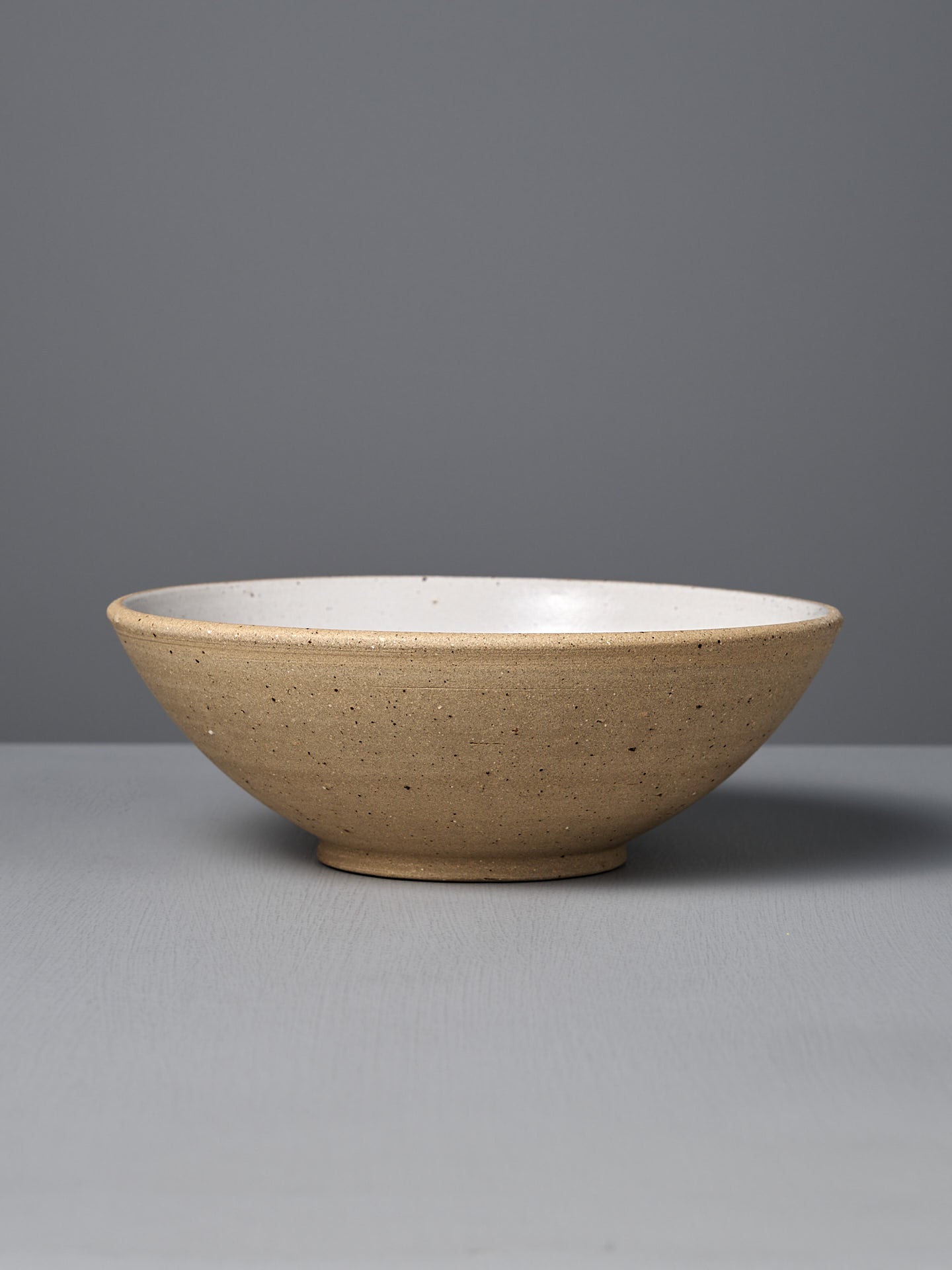 The Speckled Toast Serving Bowl – Large features a handmade ceramic design with a light brown speckled exterior and white interior, crafted in Wellington and displayed on a gray surface with a matching background.