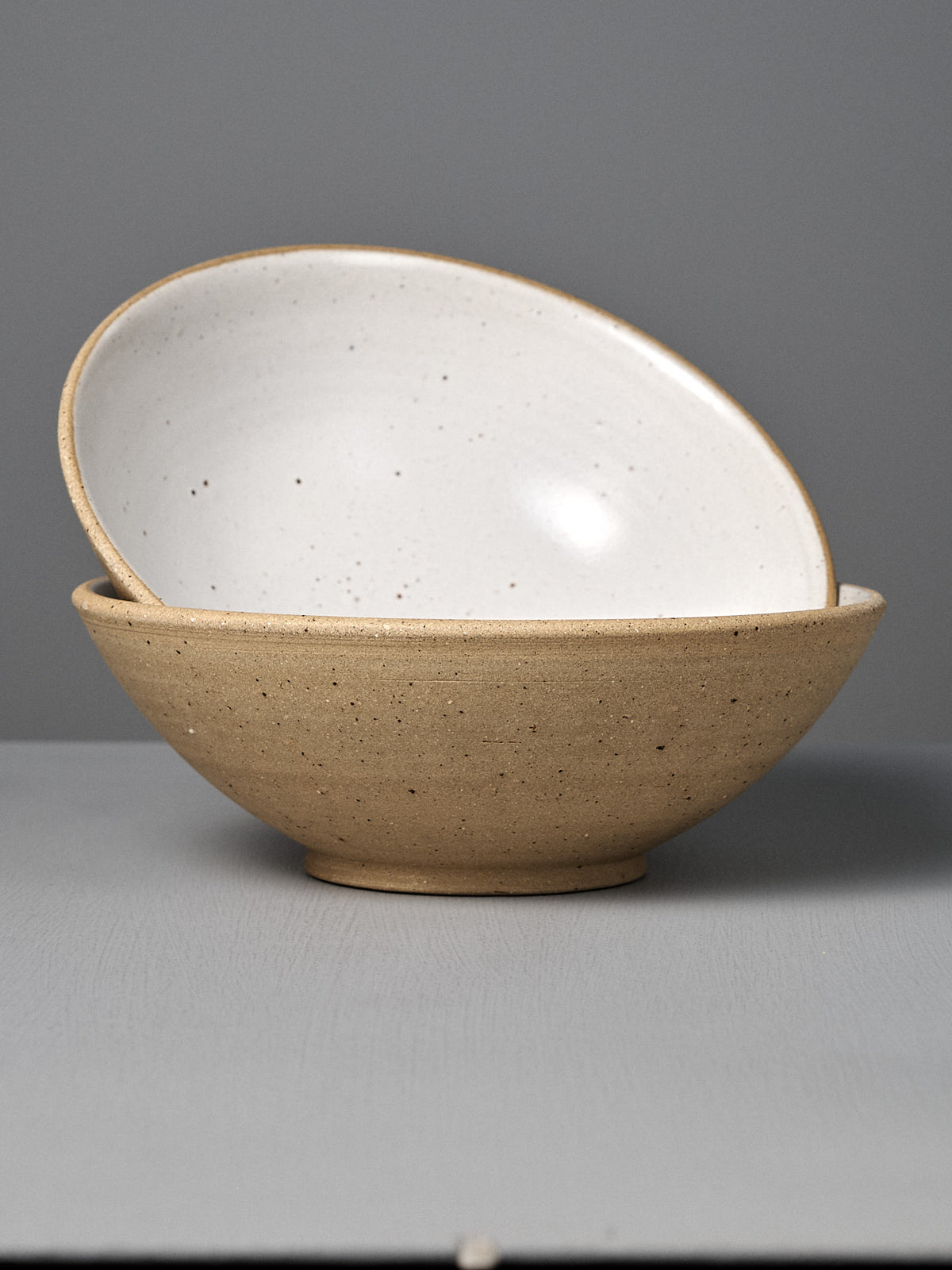 The Speckled Toast Serving Bowl – Large features a handmade design with a beige speckled exterior and white interior, shown stacked at an angle on a gray surface—crafted with care in Wellington.