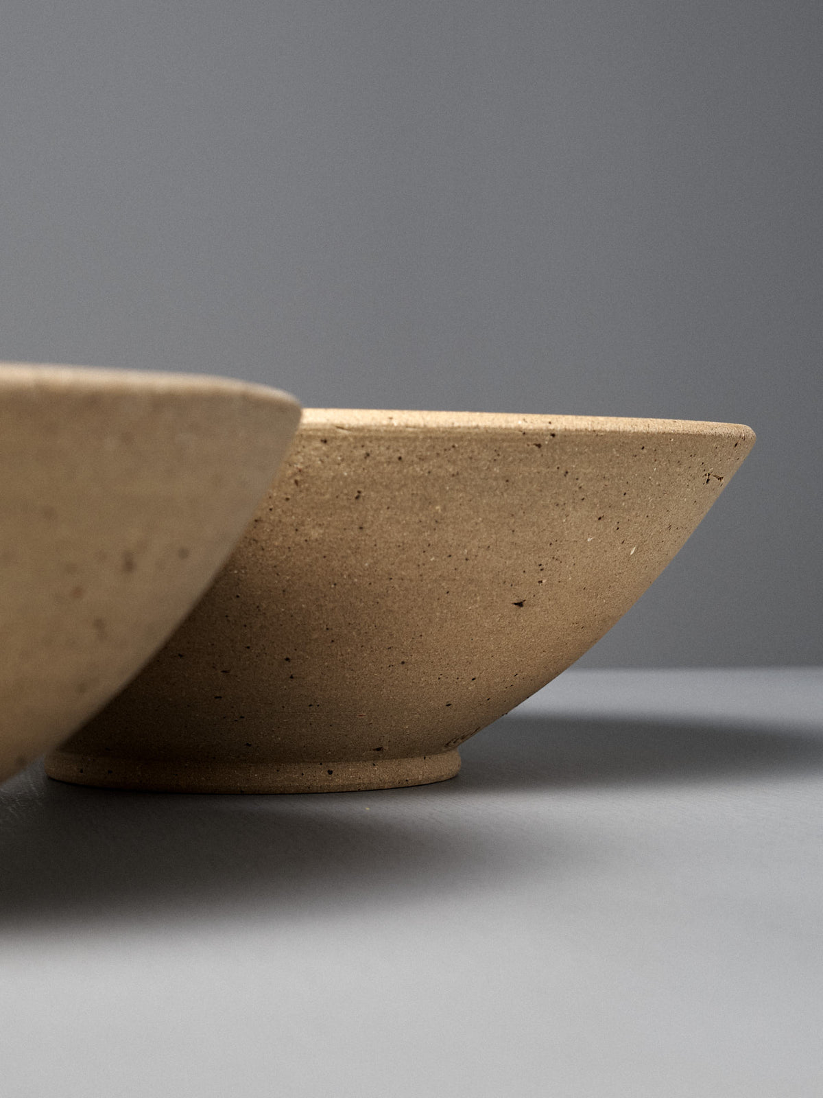 Two beige handmade ceramic bowls rest on a gray surface; one is a partially visible Speckled Toast Serving Bowl – Large in the foreground, while the other bowl is more prominently displayed.