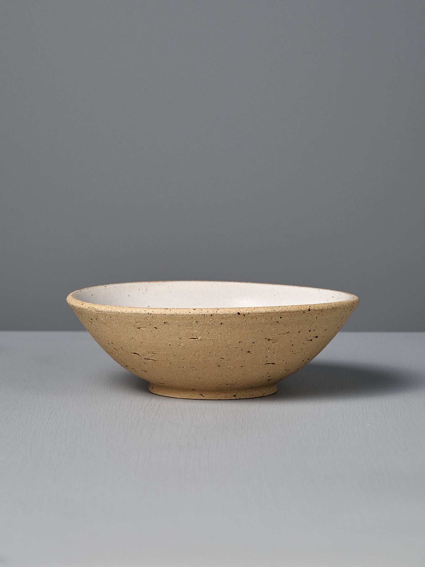The Speckled Toast Serving Bowl – Medium is a handmade, shallow, round beige ceramic bowl with a speckled texture, displayed on a light gray surface against a plain gray background—elegantly crafted in Wellington.