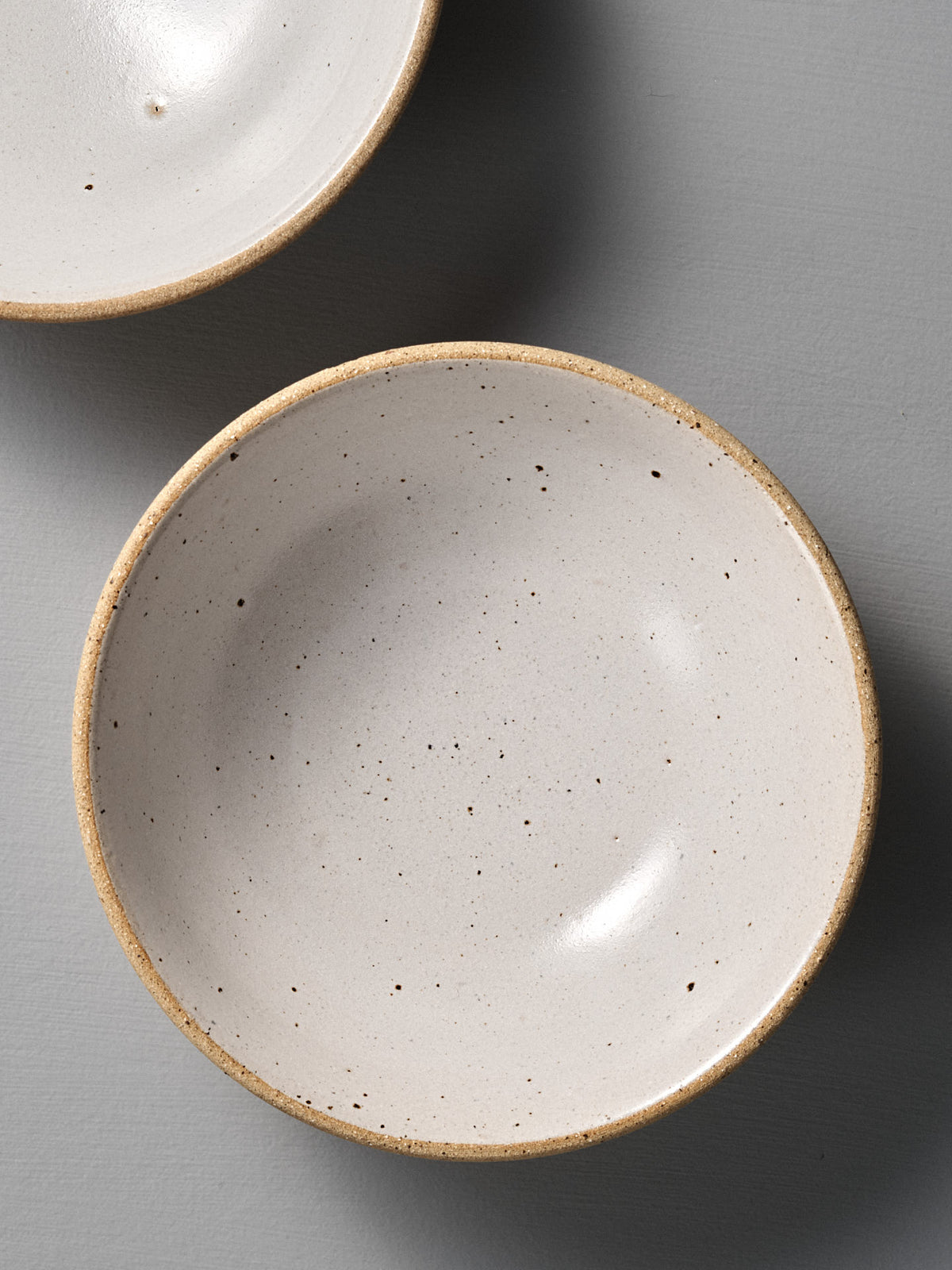 A Speckled Toast Serving Bowl – Medium with a beige rim sits on a smooth gray surface, highlighting the handmade ceramic&#39;s charm; one bowl is fully visible, while another appears partially at the top left.