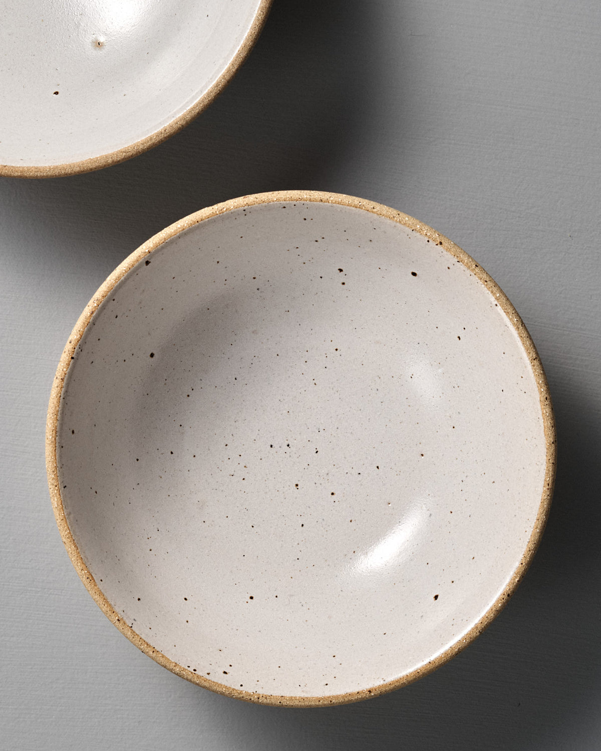 A Speckled Toast Serving Bowl – Medium with a beige rim sits on a smooth gray surface, highlighting the handmade ceramic's charm; one bowl is fully visible, while another appears partially at the top left.