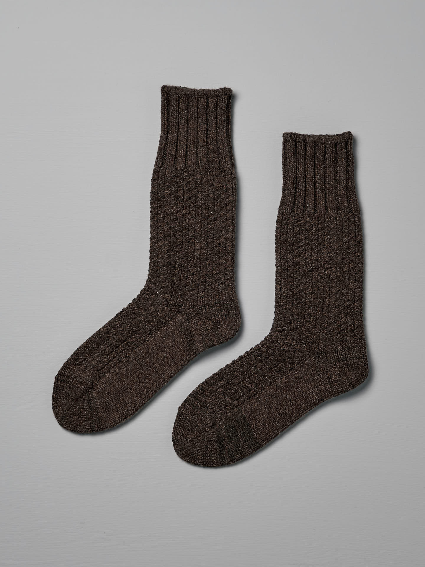 A pair of dark brown, knitted wool socks with a ribbed cuff placed side by side on a light gray background, available in both US Men's sizes and EUR sizes, **Wool Cotton Boot Socks – Brown** by **Nishiguchi Kutsushita**.