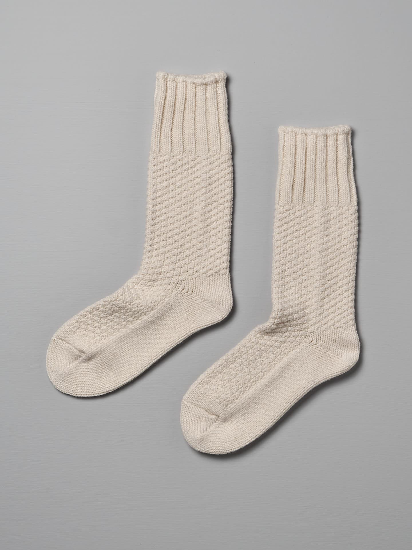 A pair of Nishiguchi Kutsushita Wool Cotton Boot Socks – Ivory with ribbed cuffs and a textured pattern on a gray background, available in Men's US and Women's US sizes.