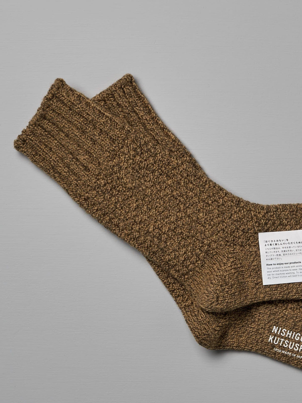 Brown, textured knit Wool Cotton Boot Socks – Mustard from Nishiguchi Kutsushita with ribbed cuffs, displayed on a gray background. There is a white label with text attached to one of the socks, indicating US Men&#39;s shoe size and EUR shoe size.