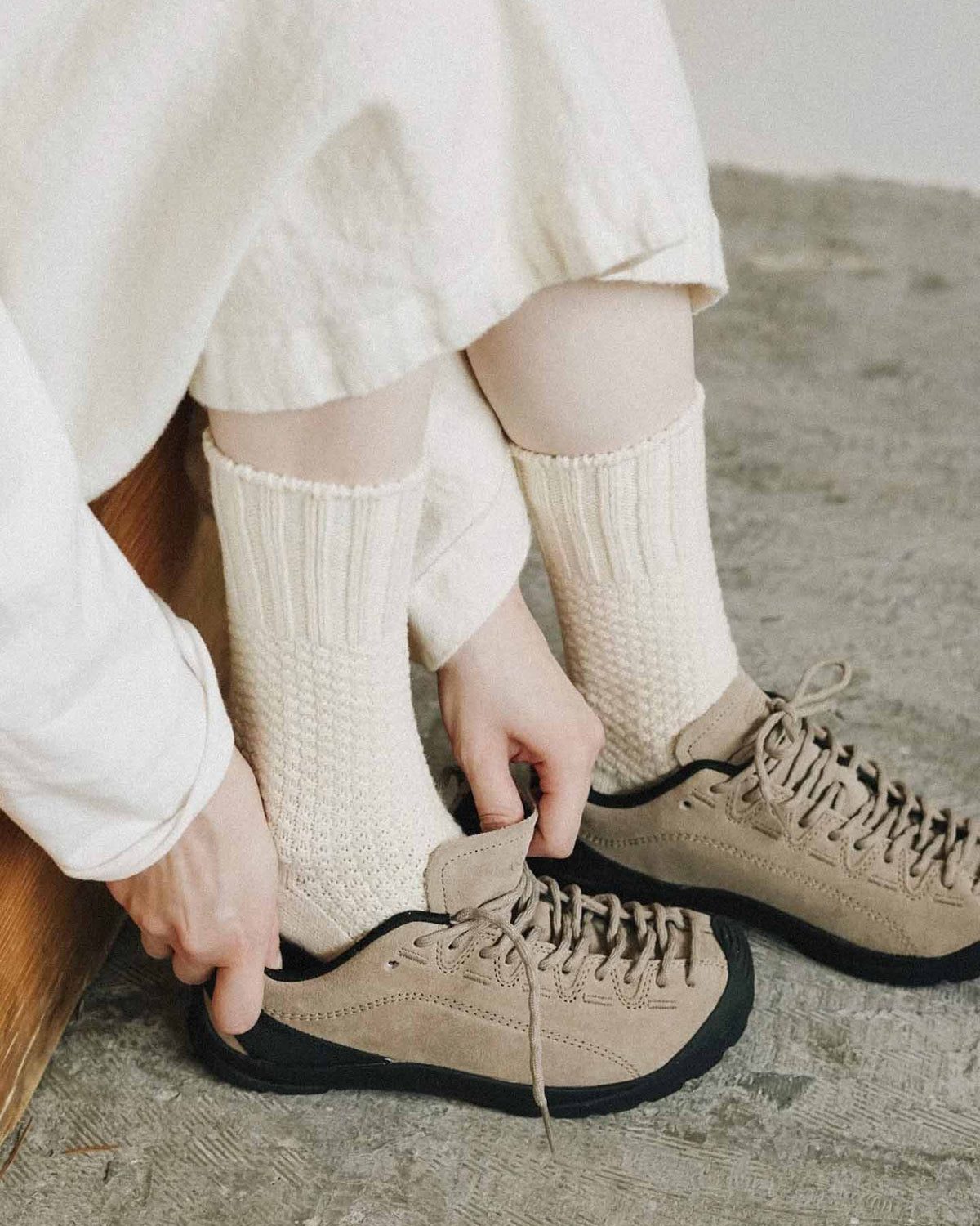 A person wearing Nishiguchi Kutsushita Wool Cotton Boot Socks – Ivory and light brown shoes, possibly consulting a size chart for men's sizing, is sitting on a wooden surface, adjusting the shoelaces of one shoe.