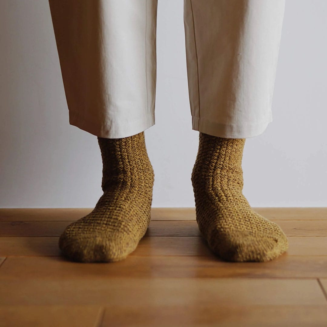 Person wearing beige pants in men&#39;s sizes and Nishiguchi Kutsushita Wool Cotton Boot Socks – Mustard standing on a wooden floor.