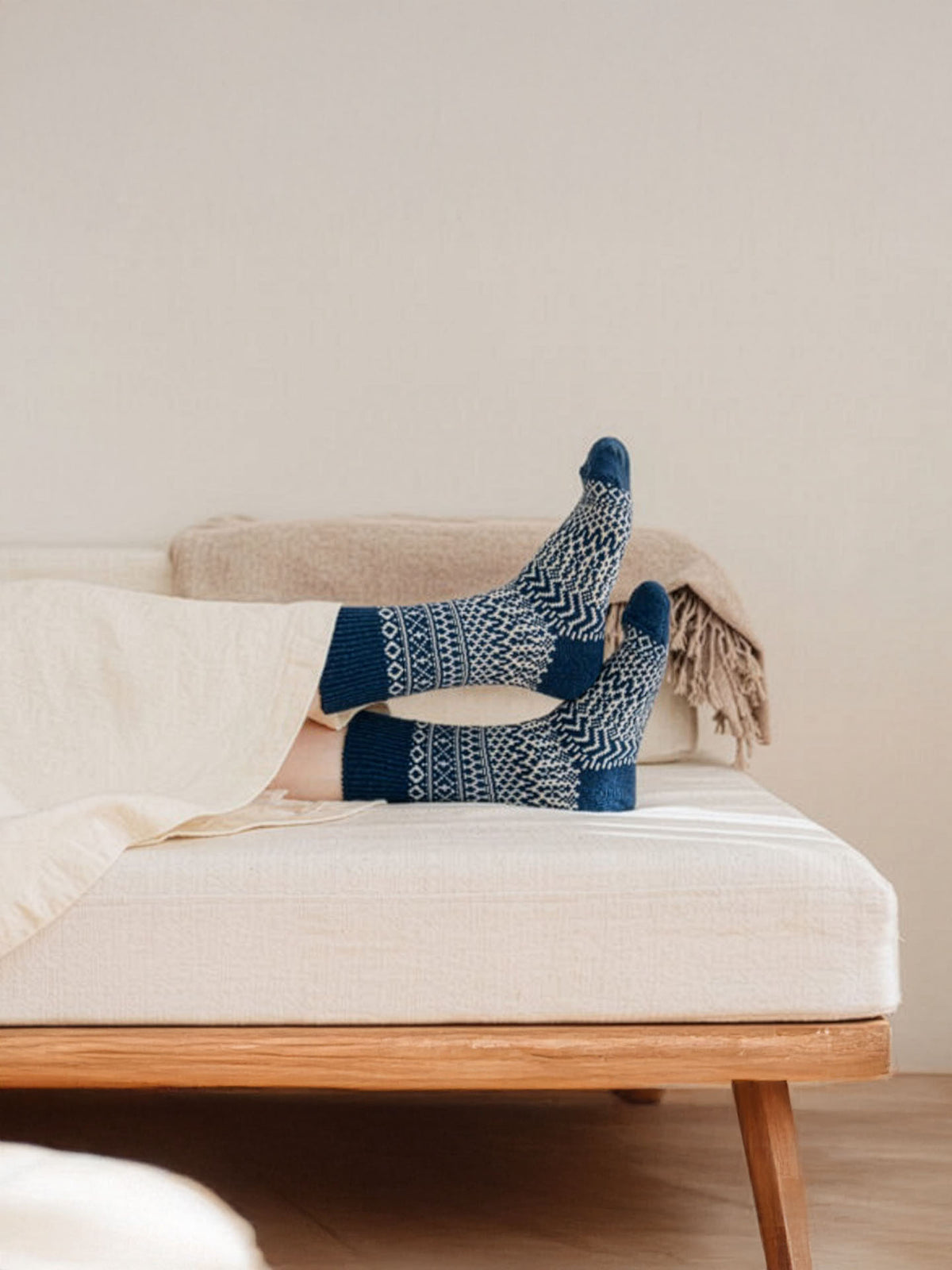 A person lies on a bed with legs crossed, wearing Oslo Wool Jacquard Socks in Seagull Blue (women's size), partially covered by a cream-colored blanket.