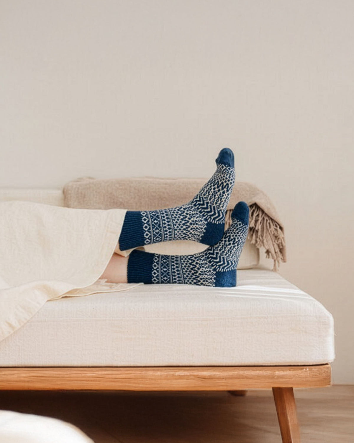 A person lies on a bed with legs crossed, wearing Oslo Wool Jacquard Socks in Seagull Blue (women's size), partially covered by a cream-colored blanket.