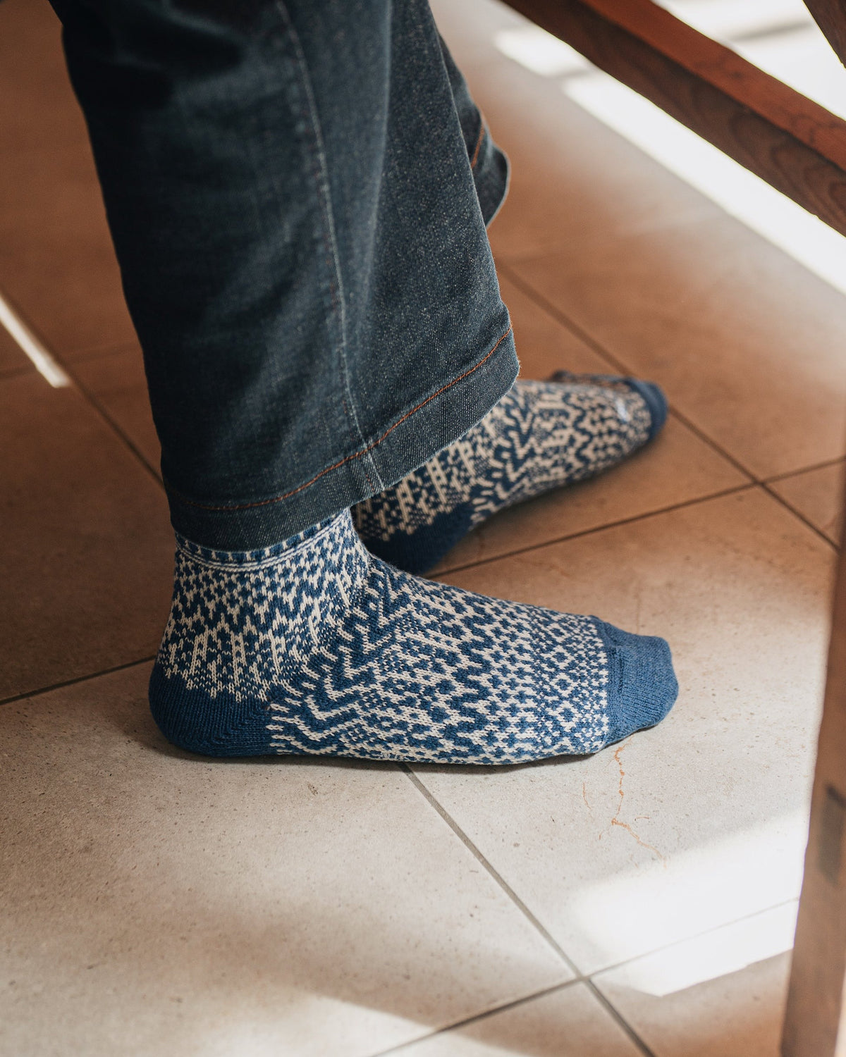 Someone wearing Oslo Wool Jacquard Socks – Seagull Blue and dark jeans stands on a tiled floor beside a wooden chair, possibly thinking about men's shoe size choices.