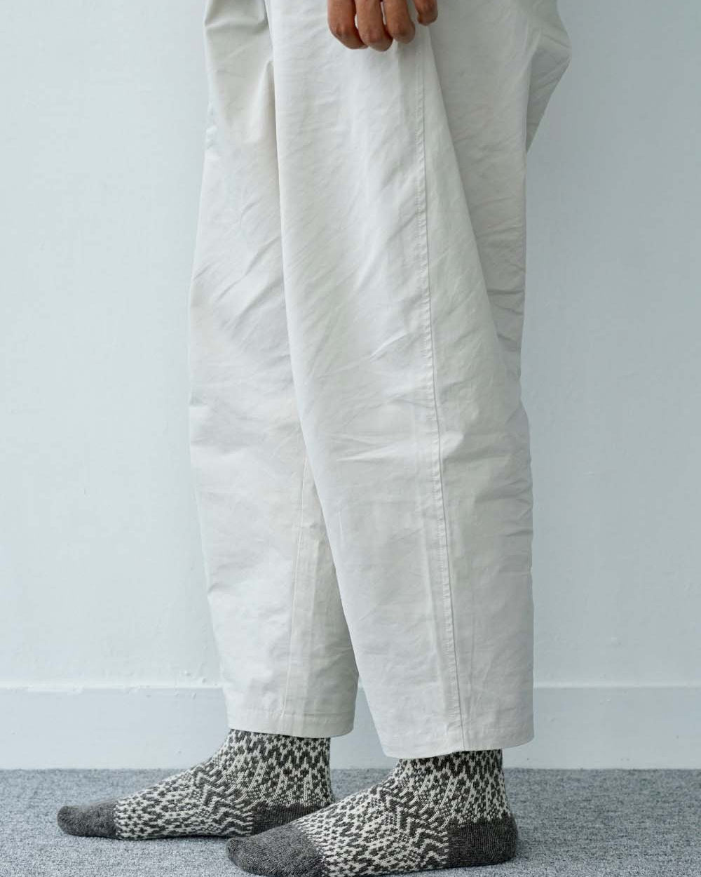 A person stands on a carpeted floor wearing loose, off-white pants and Oslo Wool Jacquard Socks – Grey; only their lower legs and one hand are visible.