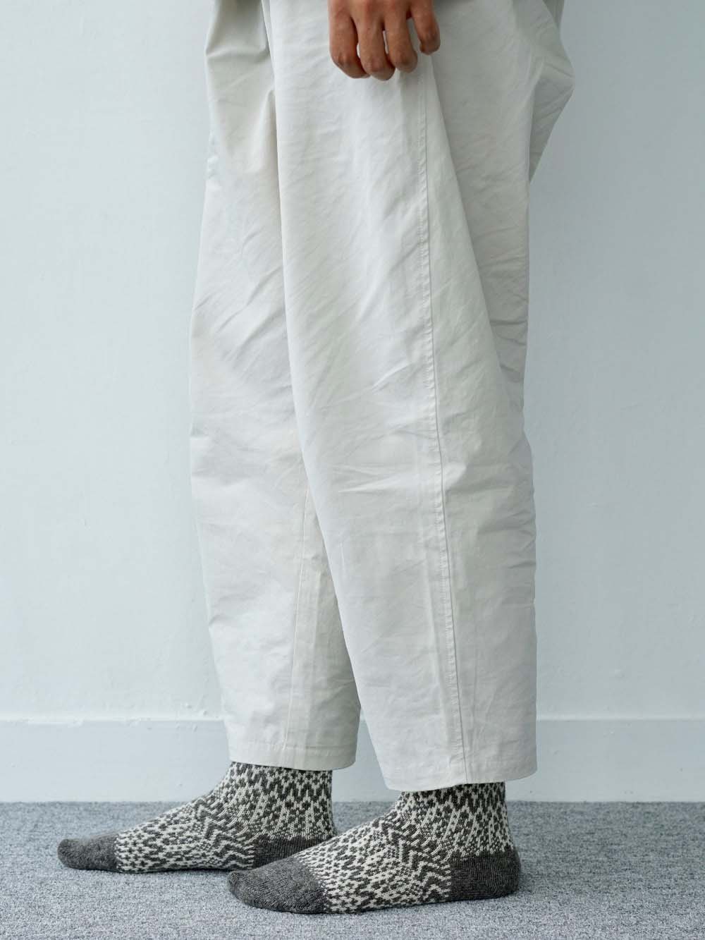 A person stands on a carpeted floor wearing loose, off-white pants and Oslo Wool Jacquard Socks – Grey; only their lower legs and one hand are visible.