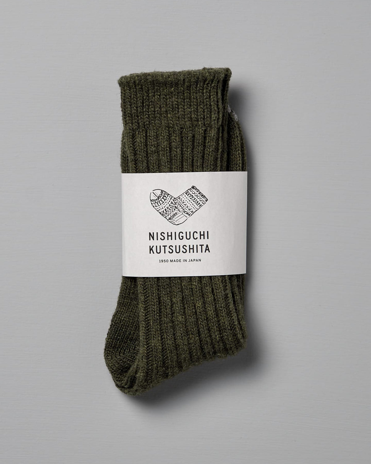 A pair of khaki wool ribbed socks folded and wrapped with a label that reads "Nishiguchi Kutsushita," with text "1985 Made in Japan." Includes a size chart for Men's US and Women's US.