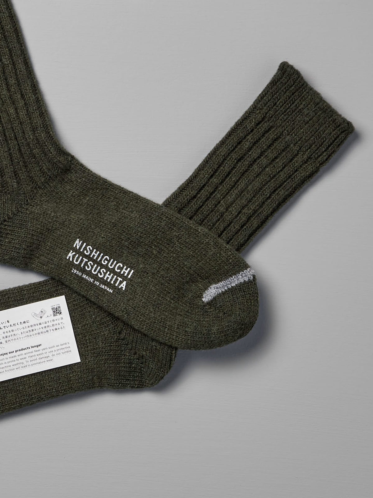 A pair of Praha Wool Ribbed Socks – Khaki with the brand name &quot;Nishiguchi Kutsushita&quot; on one sock, laid out on a gray surface. A white product label is attached to the socks, and a size chart for both Women&#39;s US and Men&#39;s US sizes accompanies them.