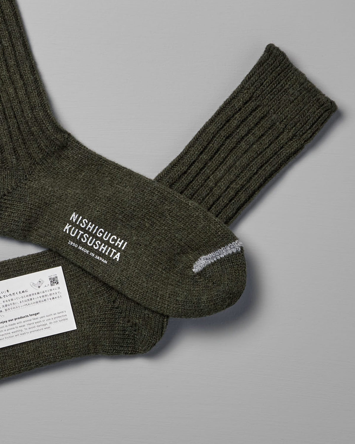 A pair of Praha Wool Ribbed Socks – Khaki with the brand name "Nishiguchi Kutsushita" on one sock, laid out on a gray surface. A white product label is attached to the socks, and a size chart for both Women's US and Men's US sizes accompanies them.