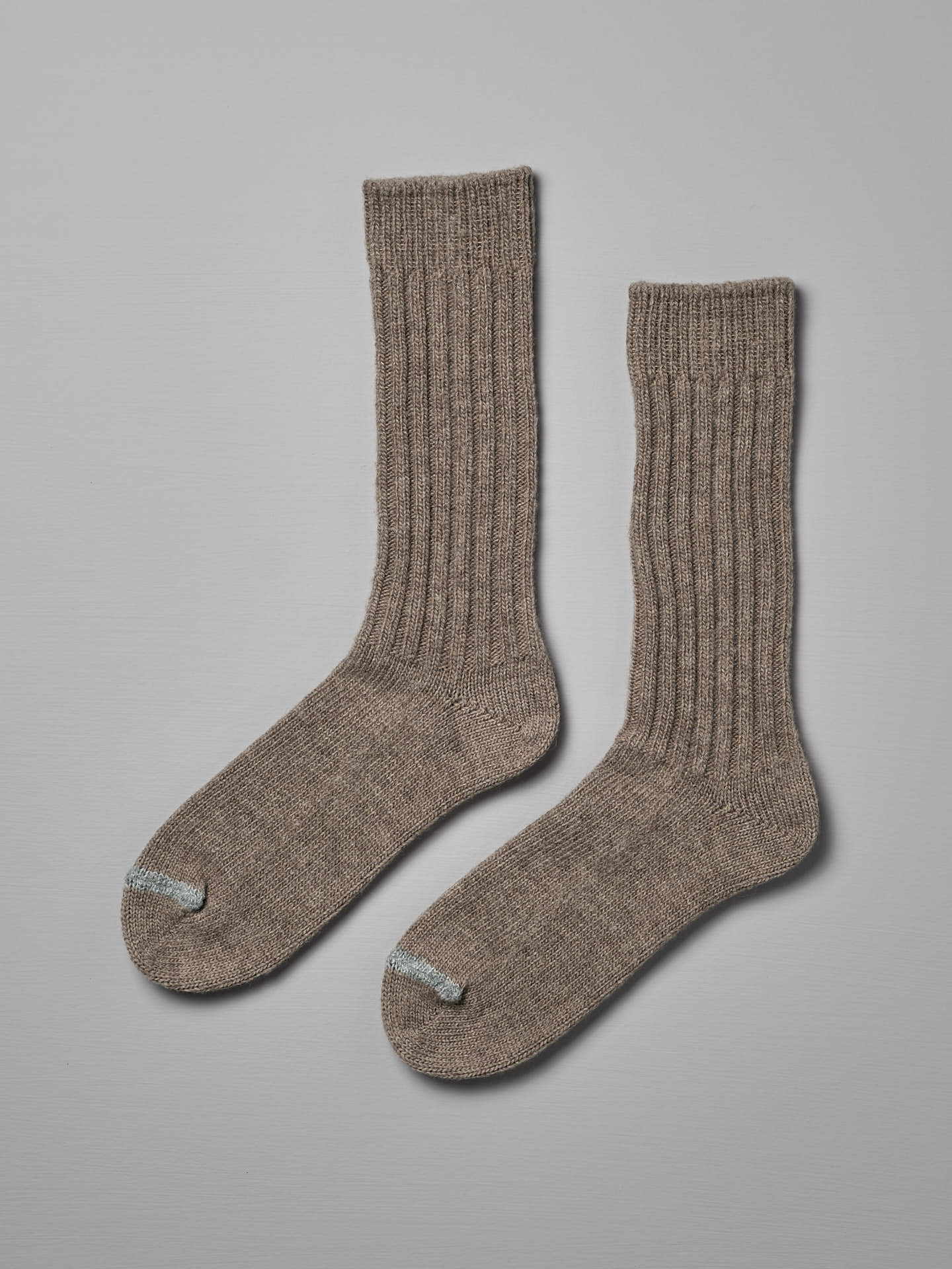 A pair of Praha Wool Ribbed Socks – Beige by Nishiguchi Kutsushita, displayed flat against a plain grey background. Both socks feature a small blue stripe near the toe area. Available in various sizes, including EUR and US (Men’s & Women's).