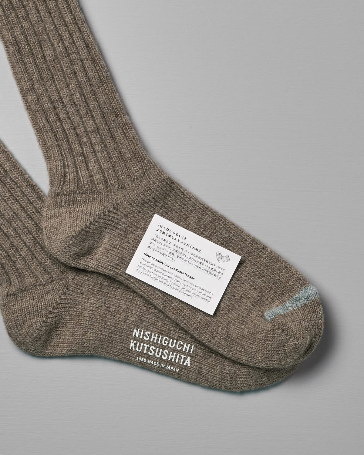 Close-up of two Praha Wool Ribbed Socks – Beige with a label featuring product care instructions, the US (Men’s) and EUR shoe sizes, and the brand name "Nishiguchi Kutsushita" printed on the bottom.