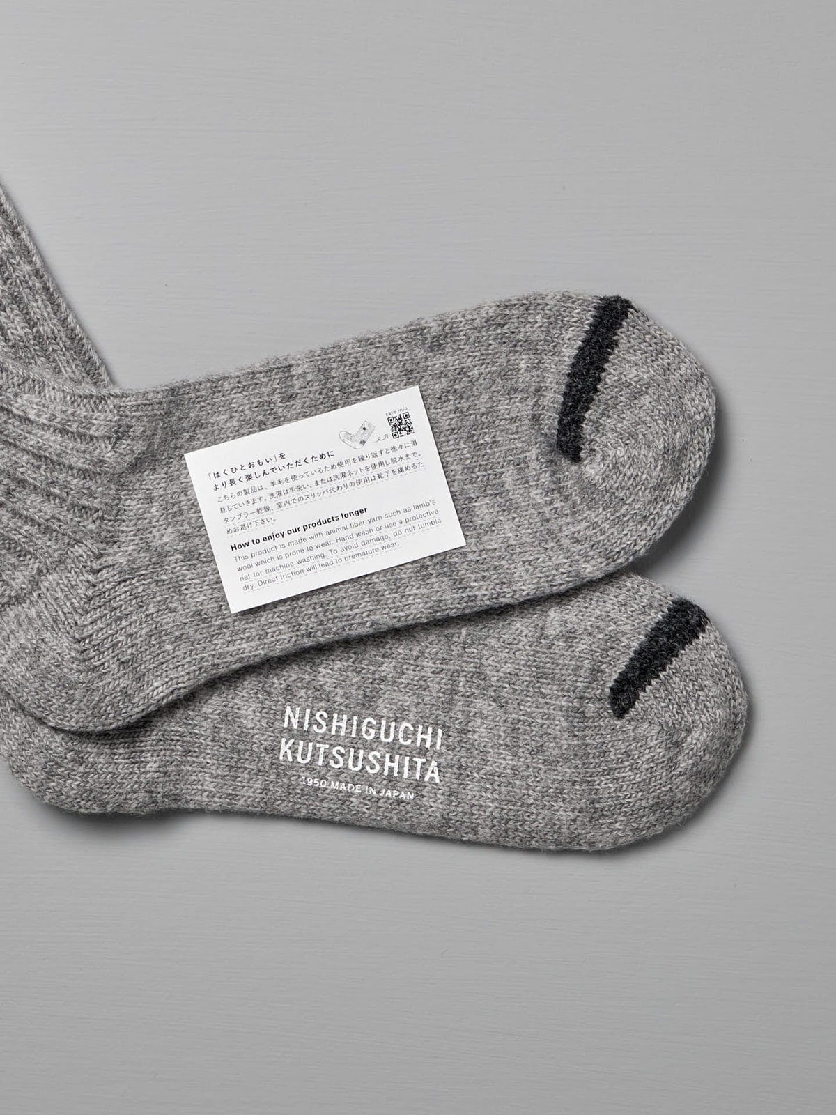 A pair of Praha Wool Ribbed Socks – Light Grey with black toe accents is laid out flat, showing the label reading &quot;Nishiguchi Kutsushita&quot; and an attached care tag with text, a QR code, and information on EUR sizes.