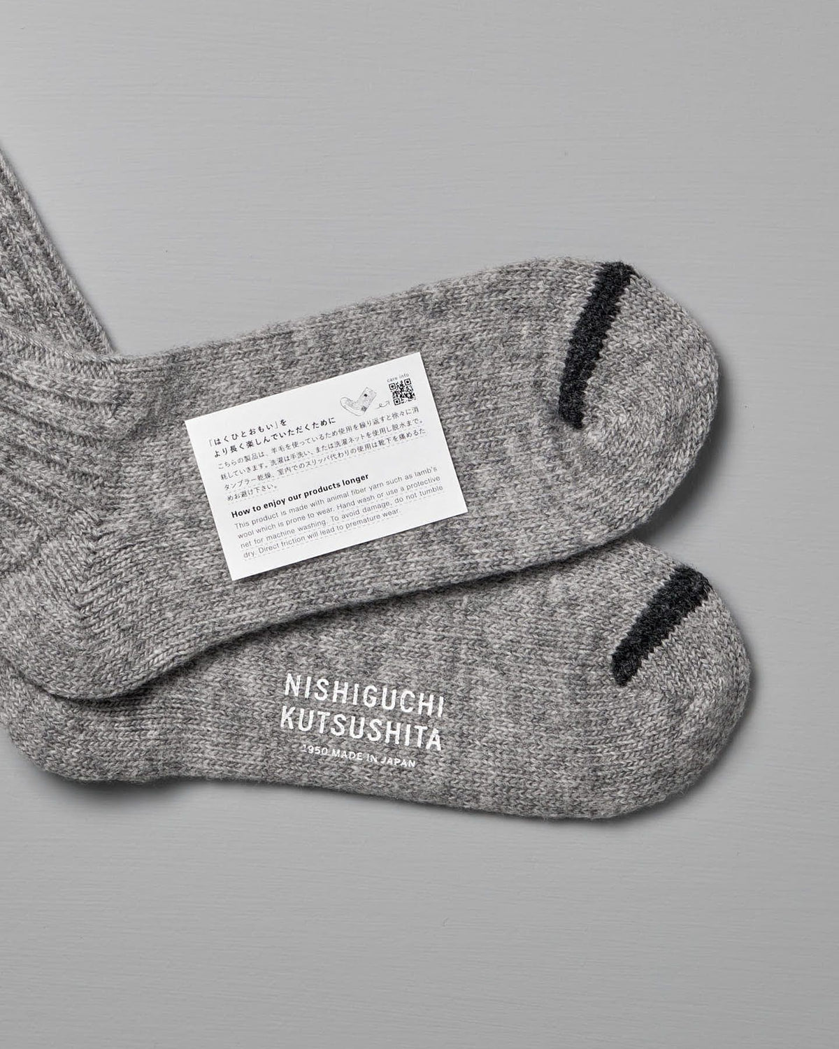 A pair of Praha Wool Ribbed Socks – Light Grey with black toe accents is laid out flat, showing the label reading "Nishiguchi Kutsushita" and an attached care tag with text, a QR code, and information on EUR sizes.