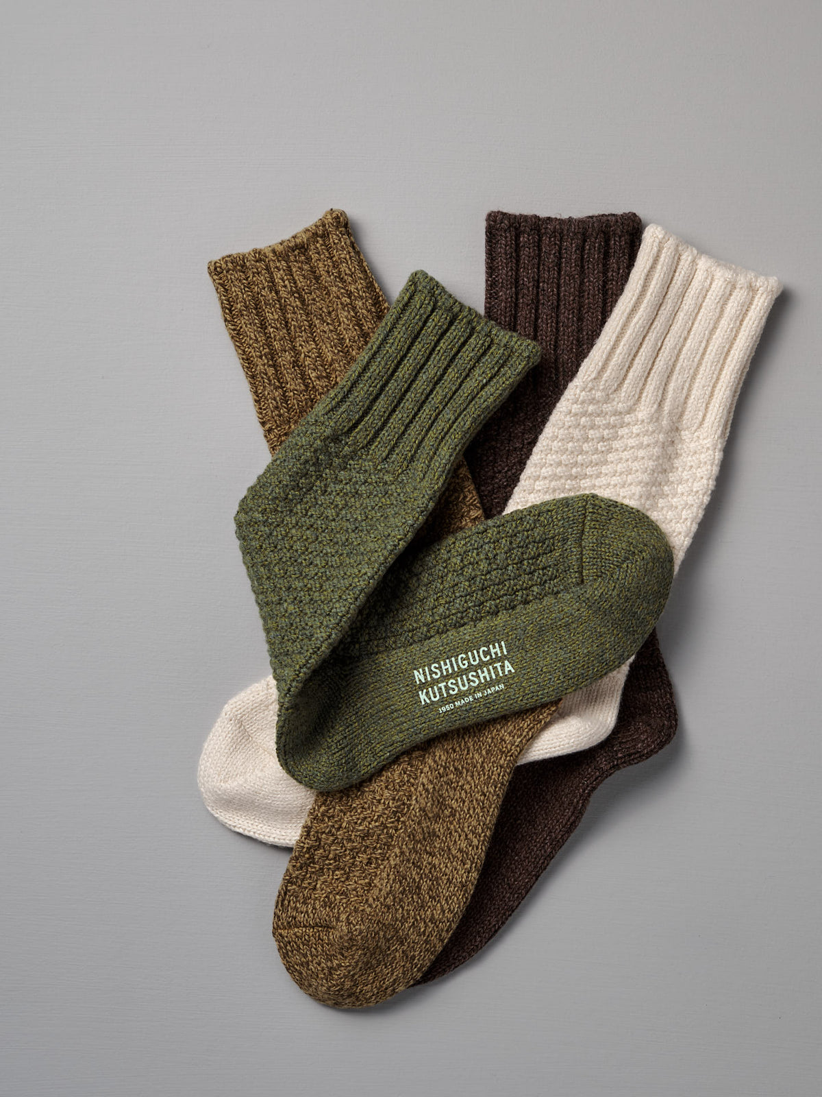 Three pairs of ribbed Wool Cotton Boot Socks – Brown by Nishiguchi Kutsushita in green, cream, and brown are displayed on a gray surface; one sock features the brand text. Refer to our women&#39;s size chart for the perfect fit.