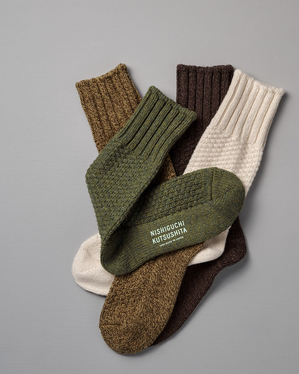 Three pairs of ribbed Wool Cotton Boot Socks – Brown by Nishiguchi Kutsushita in green, cream, and brown are displayed on a gray surface; one sock features the brand text. Refer to our women's size chart for the perfect fit.