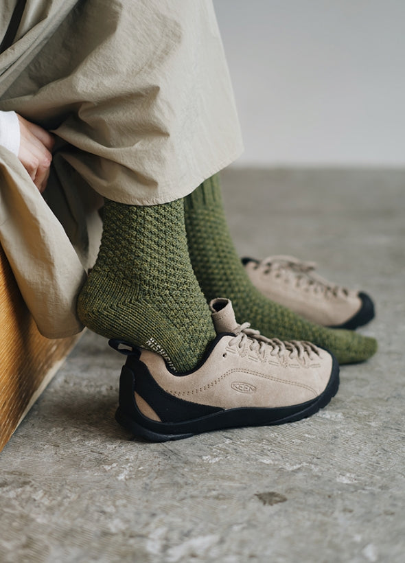 A person wearing beige pants and Nishiguchi Kutsushita Wool Cotton Boot Socks in Moss Green, along with beige sneakers (women’s US size), sits on a wooden bench on a concrete floor.