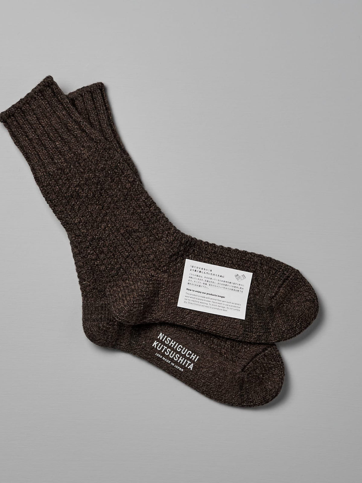 Dark brown knitted socks with a texture, laid one on top of the other against a grey background. A label on the socks shows the brand &quot;Nishiguchi Kutsushita,&quot; care instructions, and sizing information for US Men&#39;s sizes and EUR sizes. The product is Wool Cotton Boot Socks – Brown.
