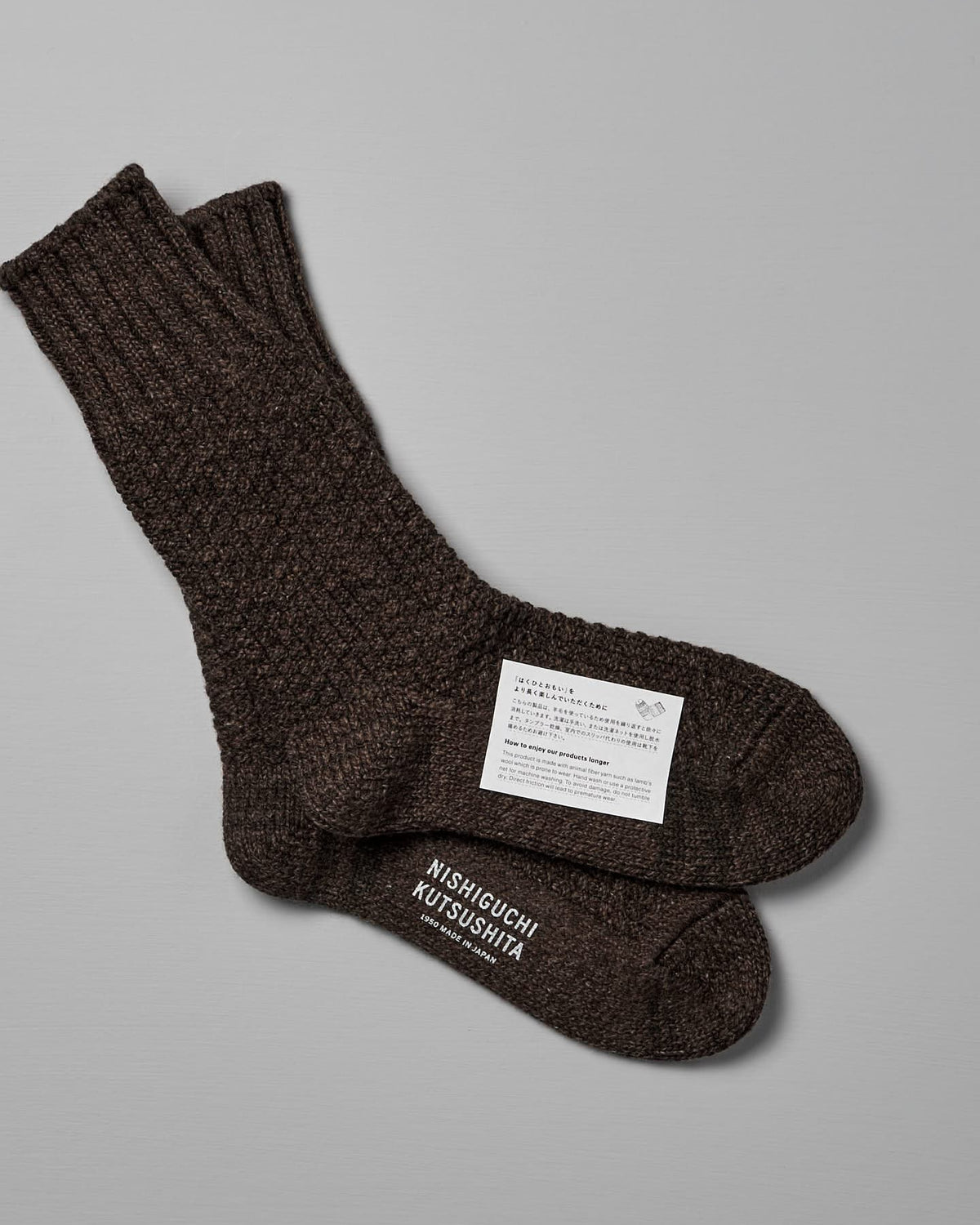 Dark brown knitted socks with a texture, laid one on top of the other against a grey background. A label on the socks shows the brand "Nishiguchi Kutsushita," care instructions, and sizing information for US Men's sizes and EUR sizes. The product is Wool Cotton Boot Socks – Brown.