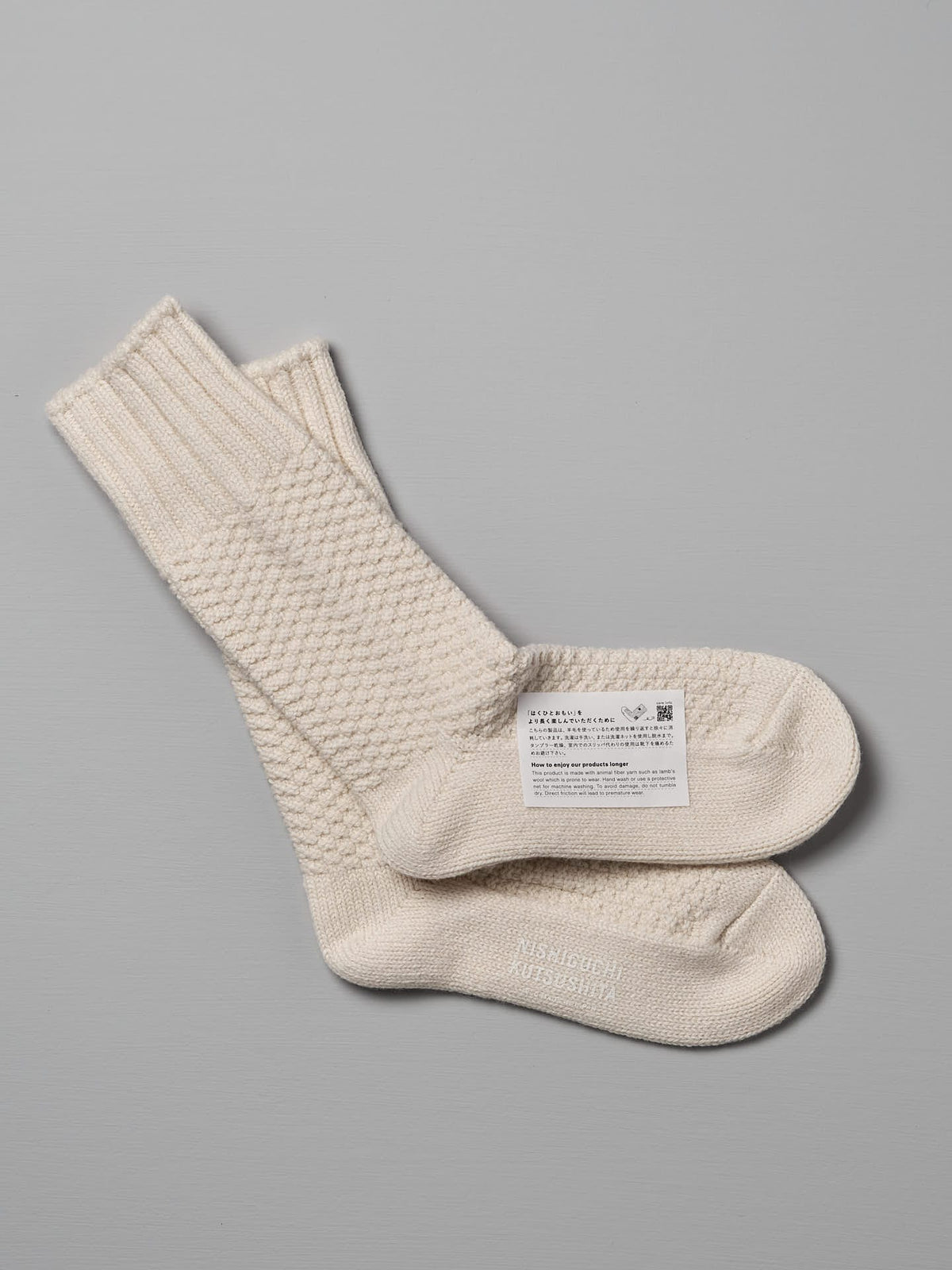 A pair of cream-colored knit socks, one folded and placed on top of the other, with a label containing text, a QR code, and size options for Women&#39;s US attached: Wool Cotton Boot Socks – Ivory by Nishiguchi Kutsushita.
