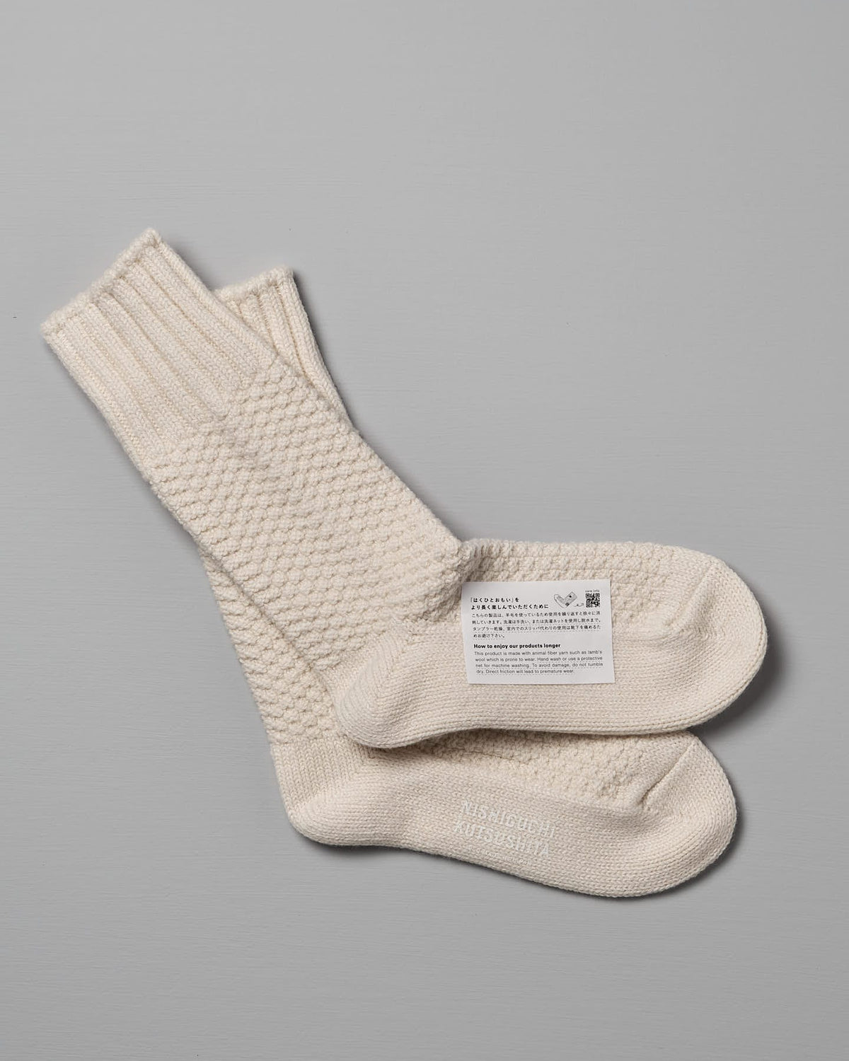 A pair of cream-colored knit socks, one folded and placed on top of the other, with a label containing text, a QR code, and size options for Women's US attached: Wool Cotton Boot Socks – Ivory by Nishiguchi Kutsushita.