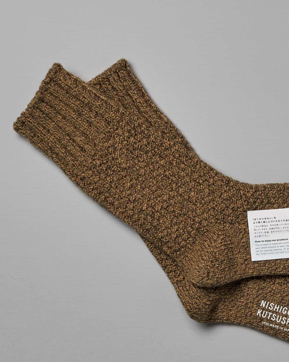 Brown, textured knit Wool Cotton Boot Socks – Mustard from Nishiguchi Kutsushita with ribbed cuffs, displayed on a gray background. There is a white label with text attached to one of the socks, indicating US Men's shoe size and EUR shoe size.