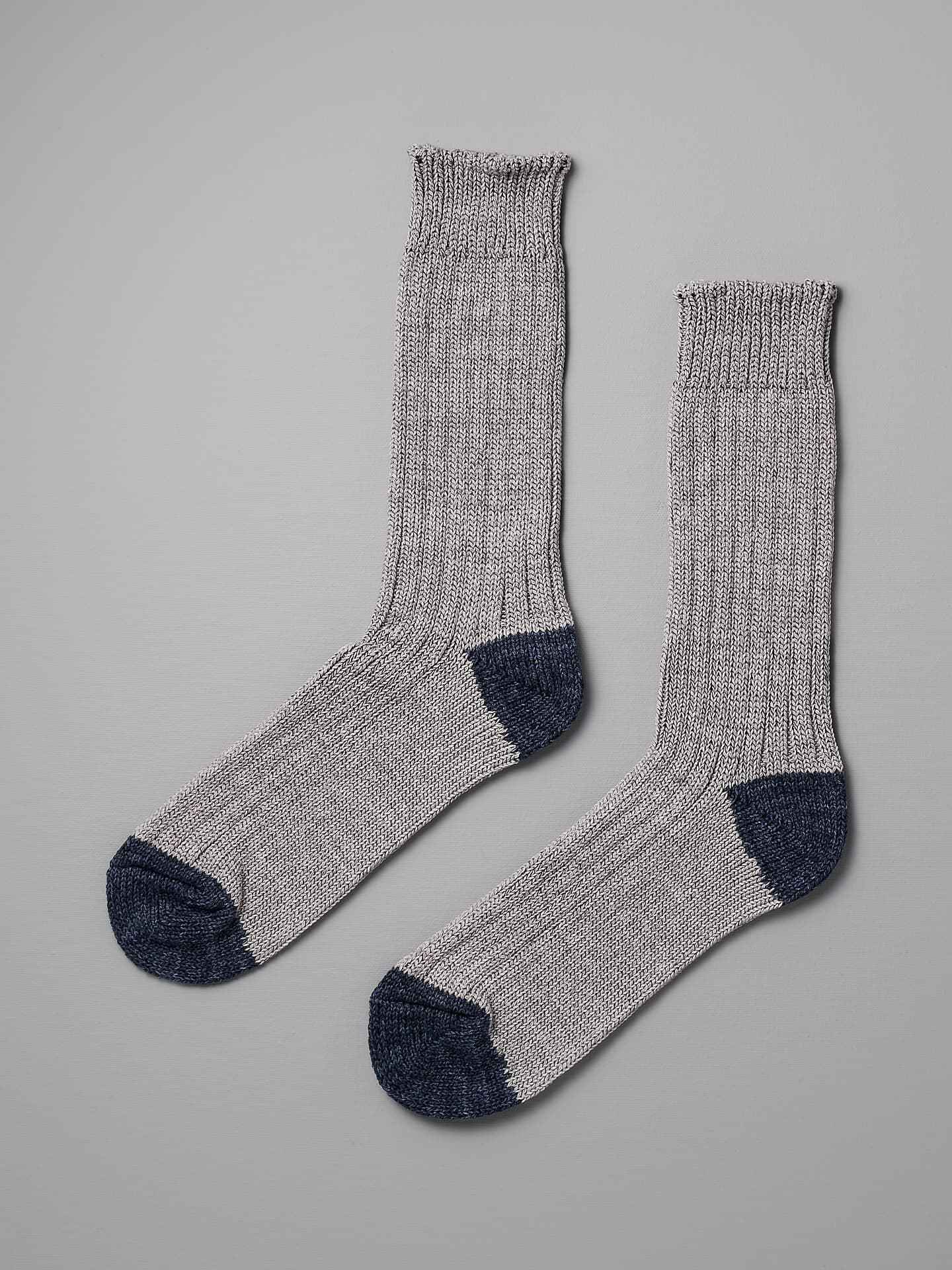 A pair of gray knitted Boston Slab Socks – Grey Denim by Nishiguchi Kutsushita with dark gray toes and heels, displayed against a plain gray background. For the perfect fit, refer to our size chart for EUR to US conversion in shoe sizes.