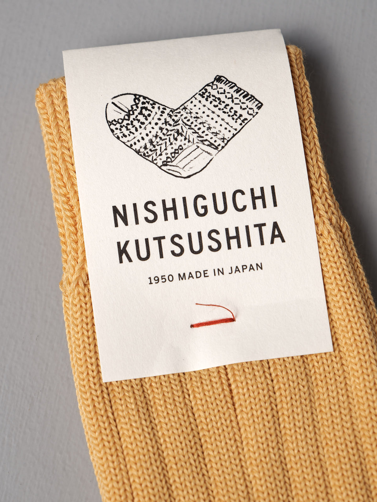Praha Cotton Socks – Susuki-Gold by Nishiguchi Kutsushita: beige knit socks with a white label featuring a sock illustration, “Nishiguchi Kutsushita 1950 Made in Japan,” and a red line. Available in women’s and men’s sizes; see size chart.