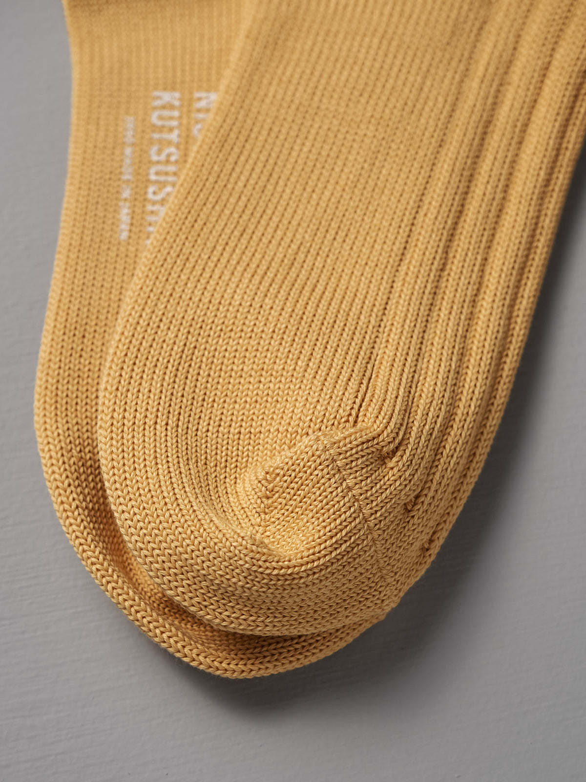 A close-up of Nishiguchi Kutsushita’s Praha Cotton Socks – Susuki-Gold, showing the mustard yellow knit and visible toe seam, displayed on a gray surface.