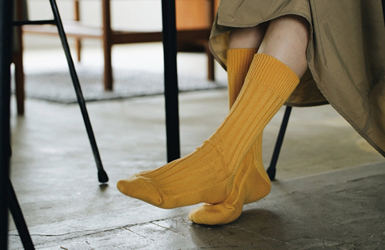 A person in a beige skirt sits indoors with crossed feet, showcasing Nishiguchi Kutsushita&#39;s Praha Cotton Socks – Susuki-Gold on a concrete floor.