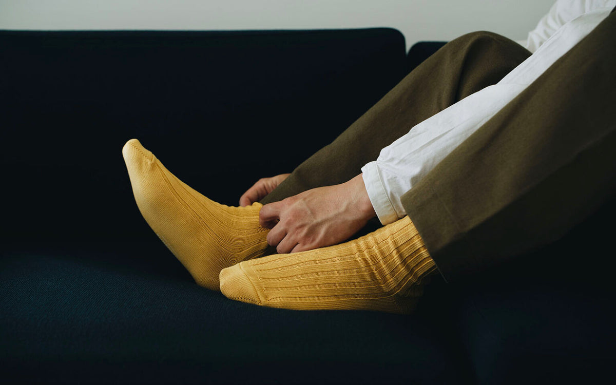 A person sits on a dark couch, wearing Nishiguchi Kutsushita’s Praha Cotton Socks – Susuki-Gold with brown pants, adjusting their socks—refer to the size chart for a perfect fit.
